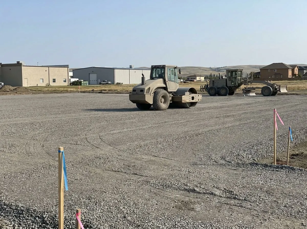 A large flat commercial building pad covered in compacted grey crushed gravel, with a smooth drum roller and a grader working across it.